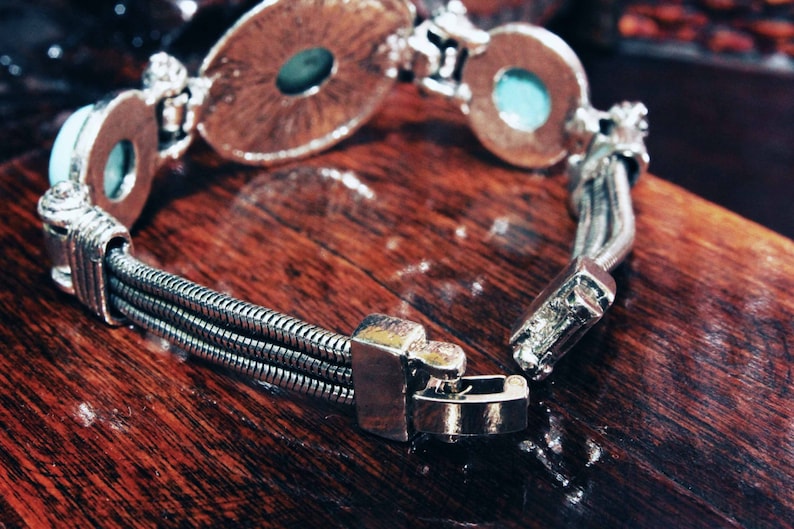 May include: A silver bracelet with three strands of chain and two round turquoise-colored stones. The bracelet has a clasp closure.