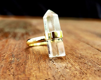 May include: A clear quartz crystal point ring with a gold band. The ring is set on a wooden surface.