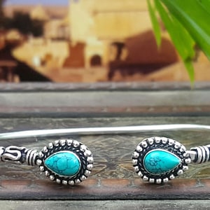 May include: Silver cuff bracelet with two turquoise teardrop-shaped stones. The stones are set in ornate, beaded bezels. The bracelet has a simple, elegant design. The bracelet is displayed on a dark, textured surface. A stylish piece of jewellery.