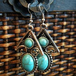 May include: A pair of silver-toned earrings with turquoise-colored oval and round stones. The earrings have a triangular top design with decorative swirls and a beaded border around the oval stone. The background is a woven brown texture.