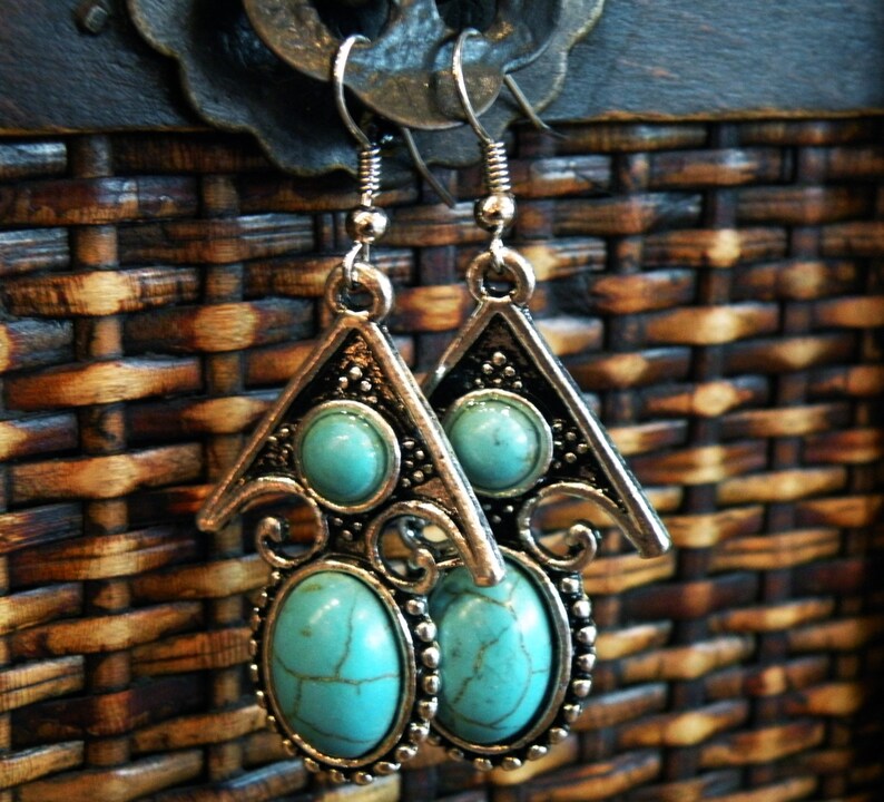 May include: A pair of silver-toned earrings with turquoise-colored oval and round stones. The earrings have a triangular top design with decorative swirls and a beaded border around the oval stone. The background is a woven brown texture.