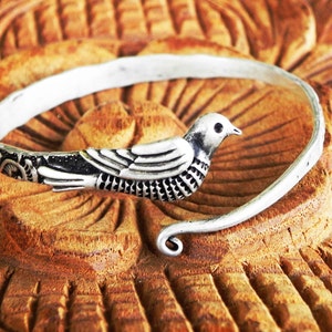 May include: A silver bangle bracelet featuring an intricate bird design. The bird is facing right and has a black eye. The bracelet is open and has a curved end.