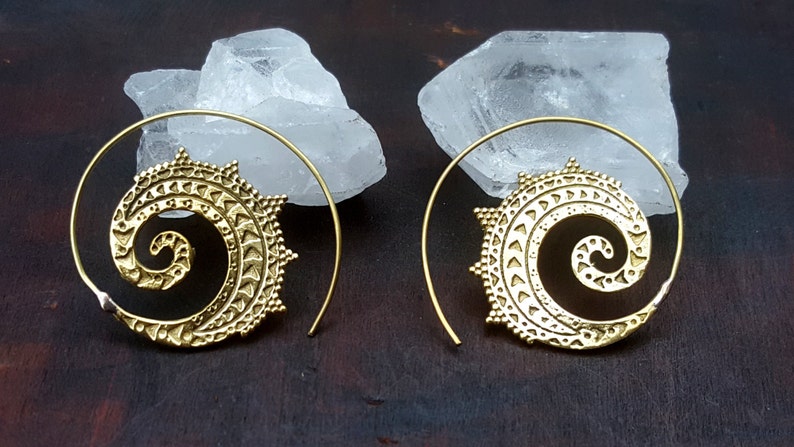May include: A pair of gold-toned spiral hoop earrings with a detailed mandala design. The earrings are made of brass and have a delicate, intricate pattern.
