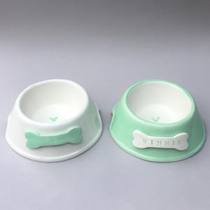 Puede incluir: Dos cuencos de cer&aacute;mica para mascotas con acabado liso. El cuenco izquierdo es blanco con un detalle en forma de hueso verde menta. El derecho es verde menta con un hueso blanco que lleva el nombre WINNIE en letras negras. Ambos tienen un coraz&oacute;n.