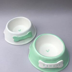 Puede incluir: Dos cuencos de cer&aacute;mica para mascotas. Un cuenco es blanco con un detalle en forma de hueso verde menta y un peque&ntilde;o coraz&oacute;n en el interior. El otro cuenco es verde menta con un detalle en forma de hueso blanco con el nombre WINNIE.
