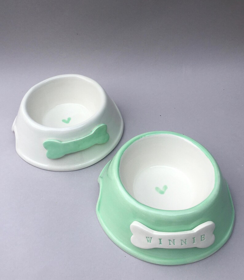 Puede incluir: Dos cuencos de cer&aacute;mica para mascotas. Un cuenco es blanco con un detalle en forma de hueso verde menta y un peque&ntilde;o coraz&oacute;n en el interior. El otro cuenco es verde menta con un detalle en forma de hueso blanco con el nombre WINNIE.