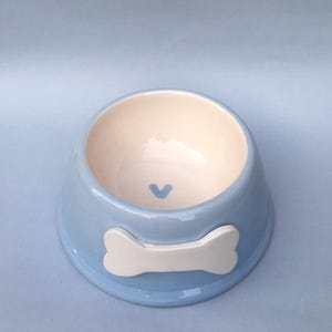 May include: A light blue ceramic pet bowl with a white bone design and a small blue heart in the center.