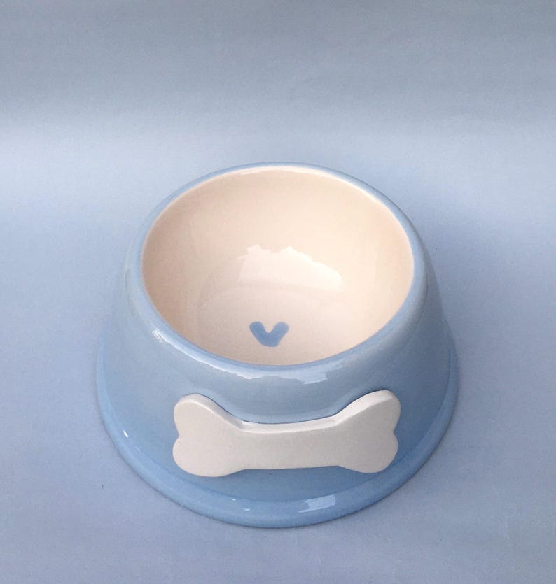 May include: A light blue ceramic pet bowl with a white bone design and a small blue heart in the center.