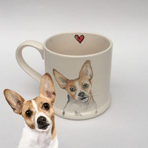 Puede incluir: Una taza de cerámica color crema con una ilustración pintada a mano de un perro marrón y blanco con orejas grandes. Un pequeño corazón rojo está pintado en el borde interior. La taza está junto a un recorte del retrato del perro.