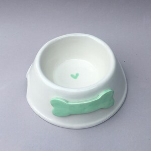 Puede incluir: Un cuenco de cer&aacute;mica blanca para mascotas con acabado brillante, que presenta un relieve en forma de hueso de color verde claro en el exterior y un peque&ntilde;o coraz&oacute;n pintado en verde claro centrado en el fondo del recipiente.