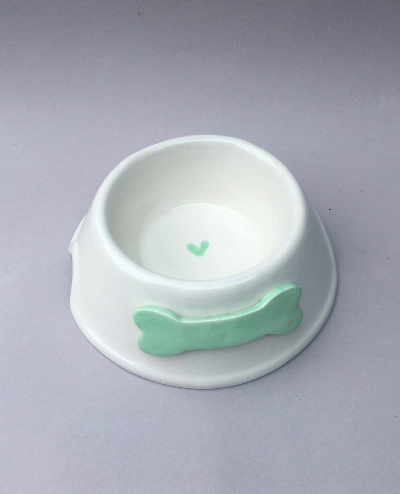 Puede incluir: Un cuenco de cer&aacute;mica blanca para mascotas con acabado brillante, que presenta un relieve en forma de hueso de color verde claro en el exterior y un peque&ntilde;o coraz&oacute;n pintado en verde claro centrado en el fondo del recipiente.