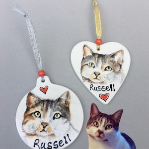 May include: Three white ceramic cat ornaments with a painted portrait of a cat. One is round, one is heart-shaped, and both have the name "Russell" written on them. A red heart is painted on each ornament. A real cat is in the image.