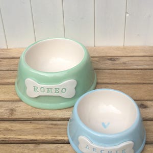 May include: Two ceramic pet bowls, one green and one blue, with bone-shaped name tags. The green bowl says "Romeo" and the blue bowl says "Archie".