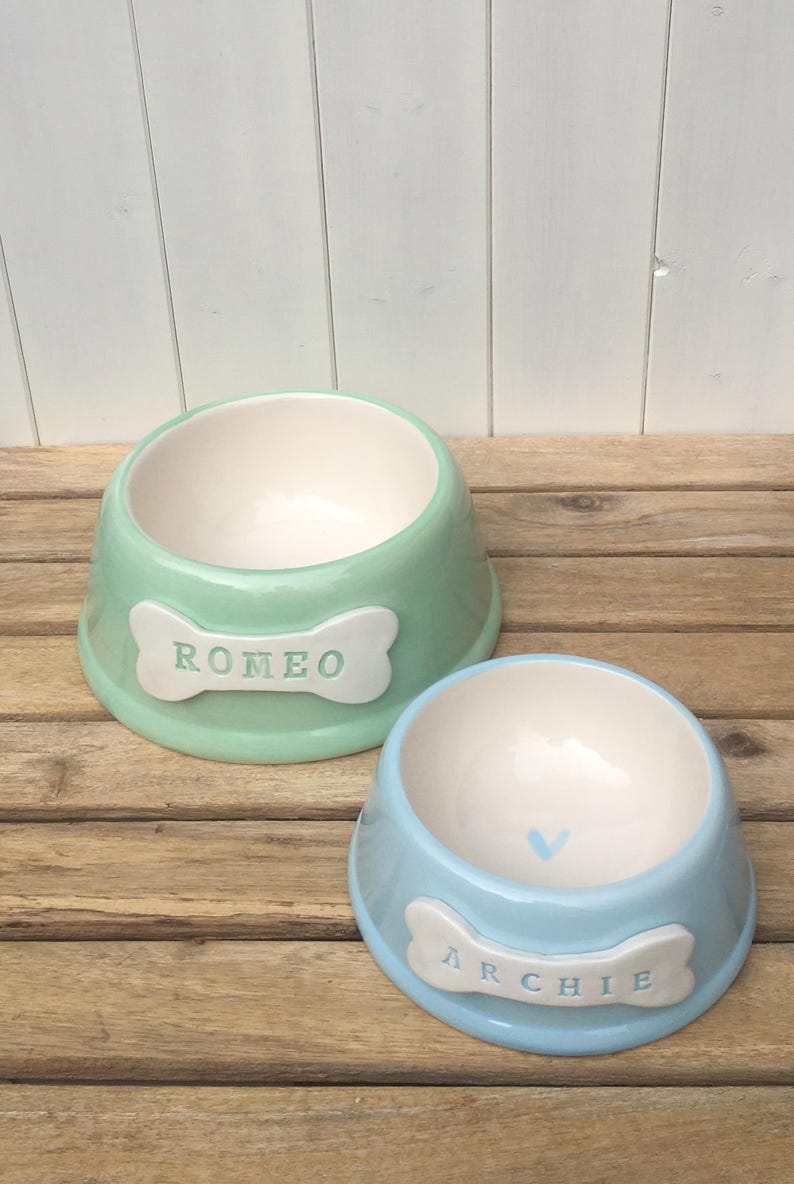 May include: Two ceramic pet bowls, one green and one blue, with bone-shaped name tags. The green bowl says "Romeo" and the blue bowl says "Archie".