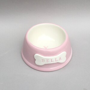 Puede incluir: Un cuenco redondo de cer&aacute;mica para mascotas en color rosa claro con un acabado liso y brillante. El interior es blanco y presenta un peque&ntilde;o coraz&oacute;n rosa en el fondo. Una placa blanca con forma de hueso lleva el nombre BELLA en letras rosas.