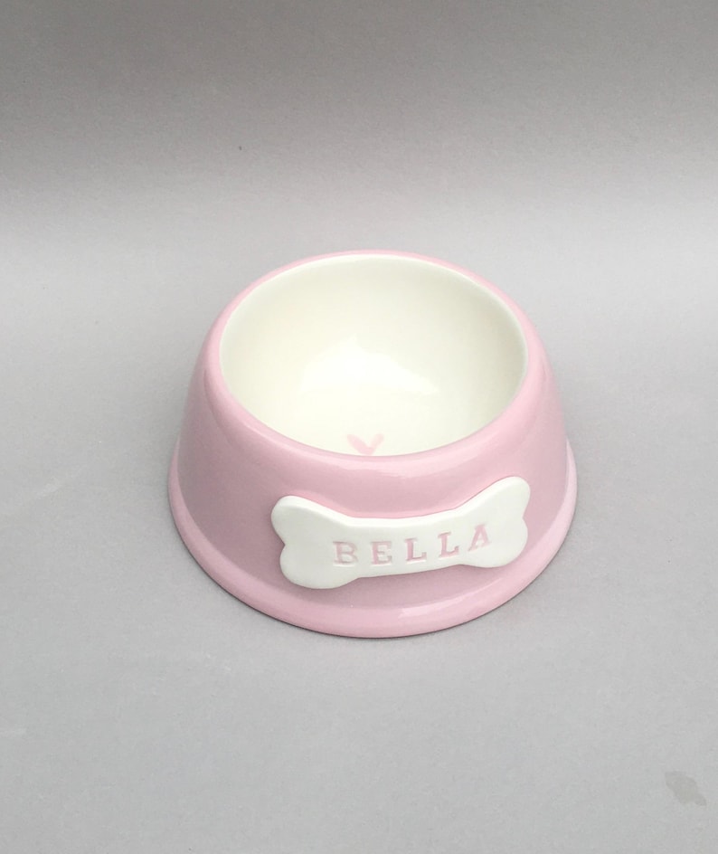 Puede incluir: Un cuenco redondo de cer&aacute;mica para mascotas en color rosa claro con un acabado liso y brillante. El interior es blanco y presenta un peque&ntilde;o coraz&oacute;n rosa en el fondo. Una placa blanca con forma de hueso lleva el nombre BELLA en letras rosas.