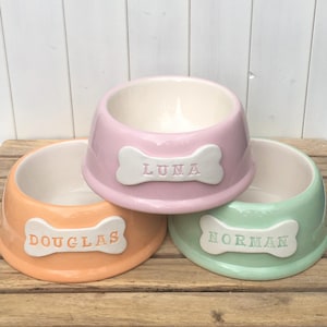 May include: Three ceramic pet bowls in pink, orange, and green with white bone-shaped name tags. The pink bowl says "LUNA", the orange bowl says "DOUGLAS", and the green bowl says "NORMAN".