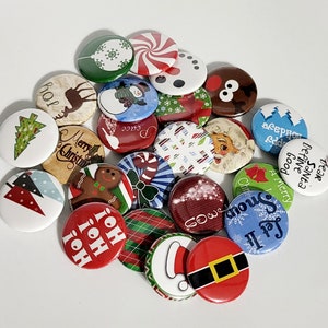 Puede incluir: Una colección de botones con temática navideña con varios diseños. Los botones presentan imágenes de Papá Noel, árboles de Navidad, muñecos de nieve y textos festivos como "Joy", "Merry Christmas" y "Let it Snow". Los botones son redondos y de varios colores.
