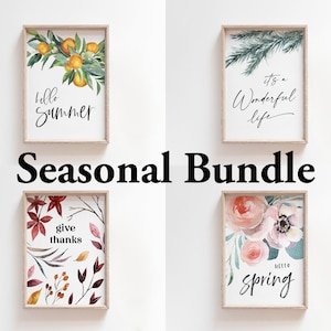 May include: A set of four printable wall art prints featuring watercolour illustrations of seasonal themes. The prints include a summer scene with oranges and leaves, an autumn scene with red and orange leaves, a winter scene with a pine branch, and a spring scene with pink and white flowers. The prints are all framed in a light wood frame and have a white background. The text on the prints reads "hello summer", "give thanks", "it's a wonderful life", and "hello spring".