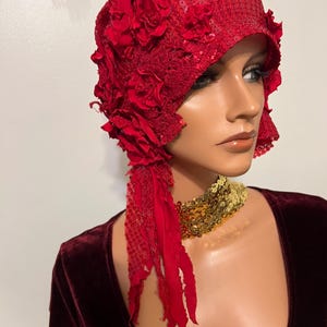 May include: A red sequined cloche hat with fabric roses and a flowing fabric accent. The hat is a vibrant red color and appears to be vintage-inspired. The hat is on a mannequin.