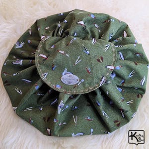 May include: A green drawstring makeup bag with a flap closure. The bag is covered in a pattern of colorful insects and other small designs. The flap has a decorative stitched border and a small, oval tag with text. The bag is open and spread out.