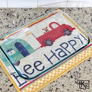 May include: A rectangular clear glass baking dish resting on a quilted fabric mat. The mat features a red truck graphic, a blue house, and the text "bee Happy!" in dark blue letters. The mat has a yellow and white gingham border trim.
