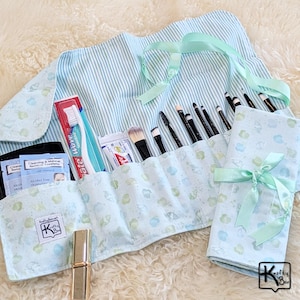 May include: A light blue and white striped makeup brush roll with a seashell pattern, holding various makeup brushes, a toothbrush, and toothpaste. A matching folded pouch is tied with a light green ribbon. The roll also contains a lipstick and cleansing towelettes.