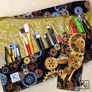 May include: A black and gold steampunk-themed makeup brush roll with various brushes, tools, and a gold ribbon. The roll features a repeating gear pattern and a small label with the text "Krafy Box".