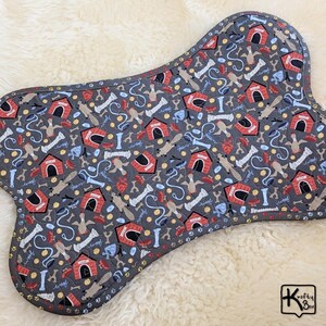 May include: Bone-shaped dog bed with a gray background and a repeating pattern of dog houses, bones, and dog silhouettes. The bed has a quilted design with a dark gray border. The word "Woof" is written in a script font throughout the pattern.