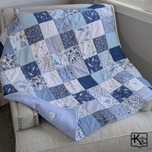 May include: A patchwork quilt featuring a grid of square fabric panels in various shades of blue, white, and grey. The patterns include clouds, moons, stars, geometric shapes, and small animals. A light blue fleece backing is visible at the corner.