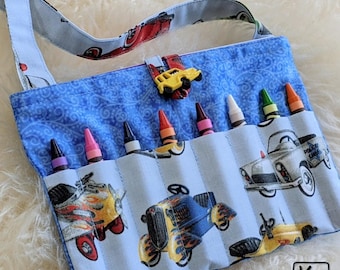 May include: Blue fabric crayon holder with a strap, featuring a car pattern. The holder has multiple pockets, each holding a crayon. A yellow car-shaped button secures the flap. The strap and flap have a car print.