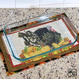 May include: A clear glass rectangular baking dish sits atop a quilted placemat featuring a turkey illustration surrounded by pumpkins and autumn leaves. The placemat is bordered by a brown fabric with a fall-themed pattern.