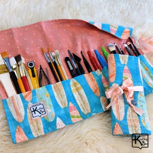 May include: A fabric art brush roll with a blue surfboard pattern and a pink polka dot interior. The roll is filled with various paint brushes, palette knives, and other art supplies. A matching roll is tied with a pink ribbon. The label says "Krafty Box".