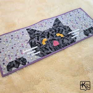 May include: A handmade, quilted table runner featuring a stylized black cat face. The cat's face is made of patterned fabric with yellow eyes and a pink nose. The background is light blue with book and script patterns, and the runner has a purple border.