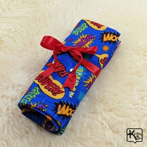 May include: A rolled-up blue fabric item with a comic book-style print, featuring words like "SUPER!" and "MOM," is tied with a red ribbon. The fabric has a dotted pattern and is decorated with star accents. The item is on a white, fluffy surface.