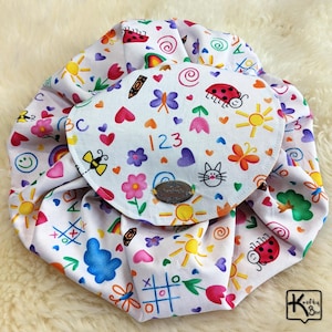 May include: A white drawstring bag with a colorful pattern of hearts, flowers, butterflies, ladybugs, suns, and the numbers 123. The bag has a flap with a metal button closure and a small logo. The bag is on a white, fluffy surface.