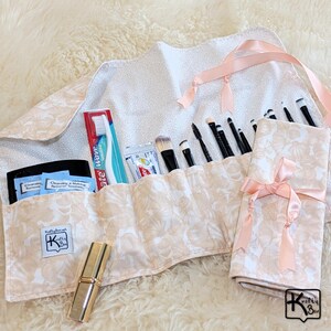 May include: A floral patterned makeup brush roll with a pink ribbon tie. The roll is filled with makeup brushes, a toothbrush, toothpaste, and other toiletries. A gold lipstick tube is also visible.