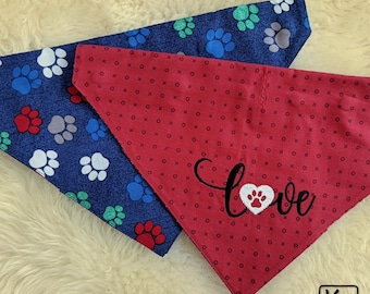 May include: Two triangular pet bandanas. One is dark blue with a repeating pattern of white, red, and green paw prints. The other is red with a repeating pattern of small black x and o symbols, featuring the word love in black script with a white paw print heart.