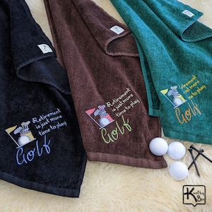 May include: Three golf towels in black, brown, and teal, each embroidered with a golf-themed design and the text "Retirement is just more time to play Golf." White golf balls and tees are also visible.