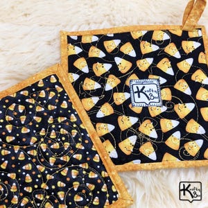 May include: Two square pot holders with a black background and a candy corn pattern. The pot holders have a gold border and a small loop for hanging. One pot holder has a label that says "Krafty Box".