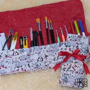 May include: A red and white dog-themed art supply roll with various art tools. The roll features a red exterior with white speckles and a dog print interior. It includes paintbrushes, crochet hooks, and other art supplies.