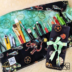May include: A black and teal steampunk-themed brush roll with various art supplies. The roll features an octopus, fish, and gears design. The roll is tied with a light green ribbon. The roll contains paint brushes, crochet hooks, and other art tools.