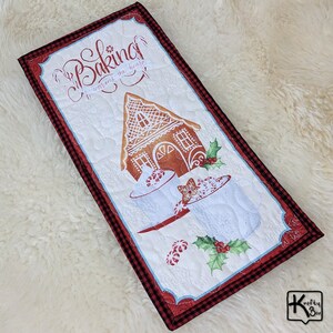 May include: A rectangular quilted fabric with a gingerbread house design. The text "Baking warms the heart" is printed at the top. The border is red and black plaid. Candy canes and holly are also featured.