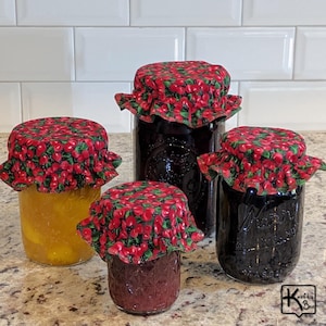 May include: Four glass jars of varying sizes, filled with preserves, are displayed. Each jar is covered with a red fabric lid patterned with cherries and green leaves. The jars contain yellow and dark red preserves.