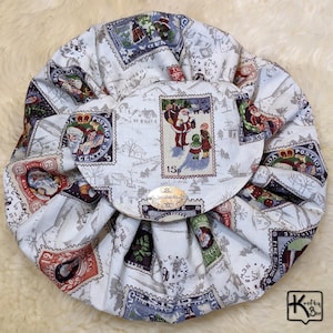 May include: A round, white drawstring bag with a Christmas stamp pattern. The bag features various postage stamp designs with festive scenes and the text "15p". The bag is gathered at the top and has a small, round flap.