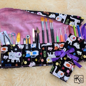 May include: A black and pink fabric roll-up case with various art and craft tools. The case features a cat pattern and the word "MEOW". The case is filled with paint brushes, crochet hooks, and other tools. A matching tag is tied with a purple ribbon.