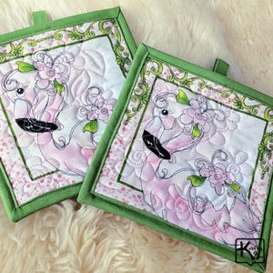 May include: Two square quilted potholders with a flamingo design. The flamingo is pink and white with a black beak and is adorned with pink flowers. The potholders have a green border and a green loop for hanging.