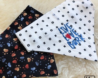 May include: Two triangular pet bandanas. One is black with a multi-coloured paw print pattern. The other is white with a black tic-tac-toe pattern and blue embroidered text that reads LIVE LOVE BARK, featuring a red heart and a paw print.