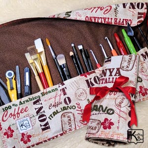 May include: A brown, rolled-up brush and tool organizer with a red ribbon. The organizer has a patterned fabric with coffee-themed text and stamps. Various art brushes, crochet hooks, and tools are visible.