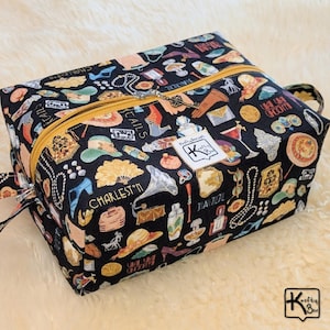 May include: A black rectangular cosmetic bag with a gold zipper and handle. The bag is covered in a pattern of vintage illustrations, including shoes, hats, and cocktails. The bag has a label that says "Krofty Box".
